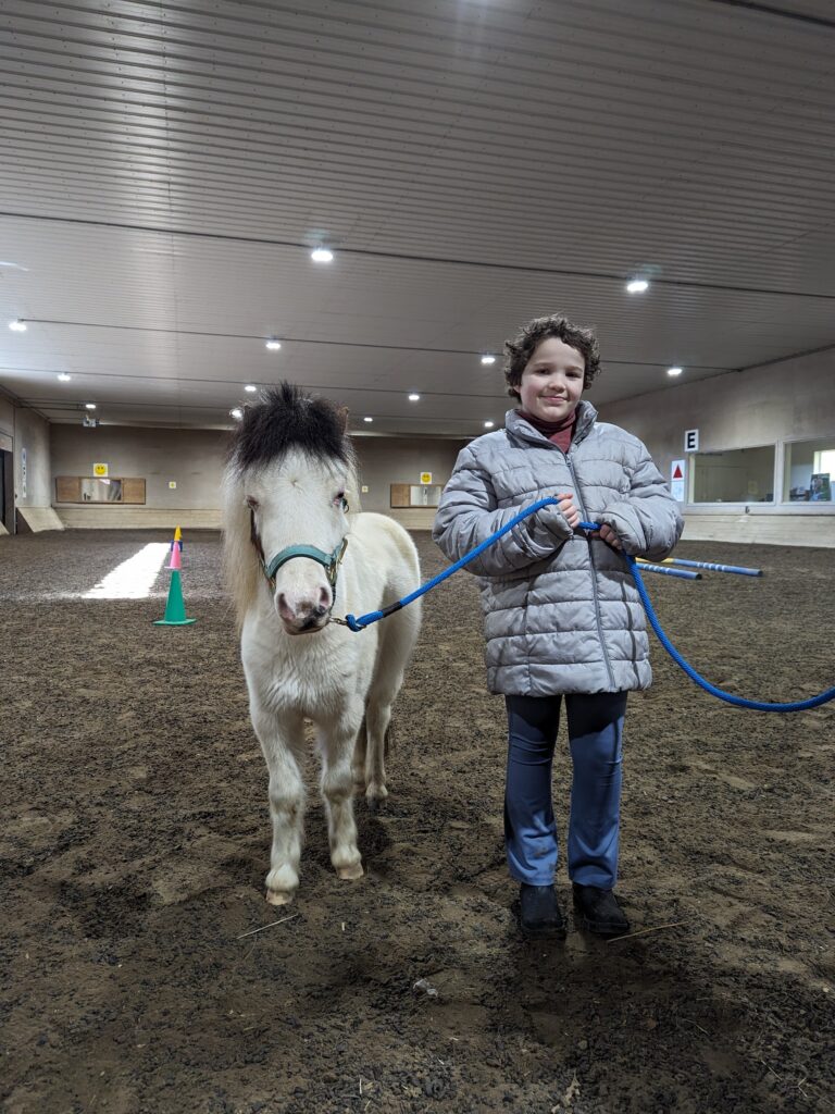 A young child smiling and holding a blue lead rope. A miniature horse named Amy stands looking curious. They are in a horse riding arena.