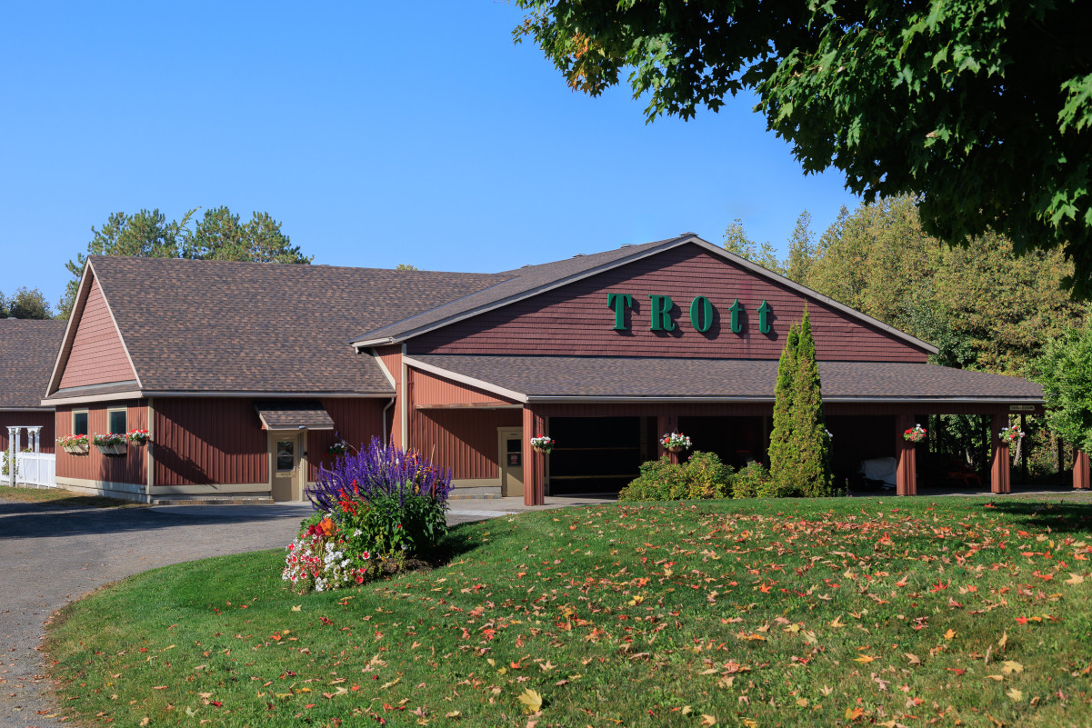 A trimmed bright green lawn with purple, red, orange, and white flowers growing fronting a large building with the words "TROtt" written across it. The premises is clean, well landscaped, and there are a few colourful fallen leaves scattered on the lawn.